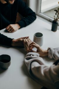 Two people holding hands across a table, offering a comforting and supportive gesture, with mugs of coffee nearby in a cozy setting.