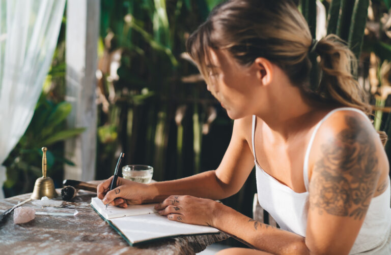 A woman sitting at a table outdoors, writing in a notebook, surrounded by crystals and a glass of water, with greenery in the background.